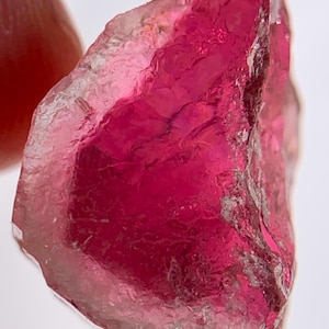 May include: A raw, translucent gemstone with a deep magenta and fuchsia color. The stone features an irregular, jagged shape with natural facets and internal textures that catch the light, displaying varying degrees of clarity and saturation.