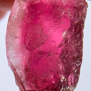 May include: A raw, translucent gemstone with a deep magenta and hot pink coloration. The stone features a natural, irregular crystalline structure with visible internal fractures, facets, and a glossy, reflective surface texture throughout.