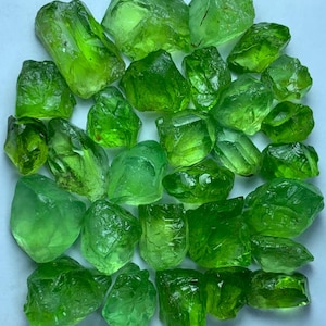 May include: A collection of raw, translucent peridot gemstones in various shades of vibrant lime and forest green. These natural, unpolished crystals feature irregular, angular shapes with faceted surfaces that catch the light, displaying a bright, glassy texture throughout the group.