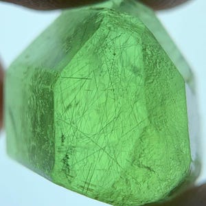 Puede incluir: Una piedra preciosa de color verde claro translúcido con forma hexagonal y superficie facetada. El interior de la piedra muestra numerosas inclusiones oscuras y finas en forma de aguja que se entrecruzan en la estructura.