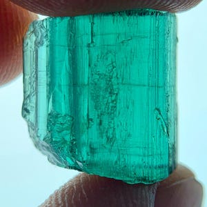 Op de afbeelding: Een levendige, doorschijnende, smaragdgroene edelsteen, vastgehouden tussen twee vingers. Het ruwe kristal heeft een rechthoekige vorm met zichtbare interne strepen en een enigszins ruw, natuurlijk oppervlak. De kleur van de edelsteen is een rijk, verzadigd groen.