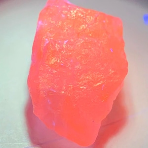 May include: A raw, unpolished mineral specimen exhibiting a vibrant, fluorescent neon pink glow. The stone has an irregular, jagged shape with a textured, crystalline surface that appears to emit light under ultraviolet illumination.