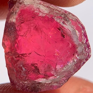 May include: A raw, unpolished gemstone with a vibrant, deep pink hue. The stone features a highly textured, irregular surface with numerous facets and internal fractures that catch the light, creating a translucent appearance with varying depth.
