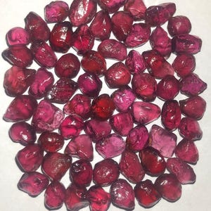 Puede incluir: Una colección de piedras preciosas de granate en bruto. Las piedras varían en tamaño y forma, con tonos rojos oscuros y rosas. Las piedras preciosas están agrupadas, mostrando su apariencia natural y sin pulir.