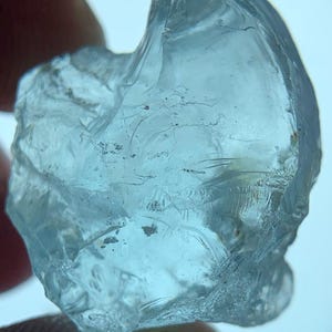 Op de afbeelding: Een ruwe, doorschijnende lichtblauwe edelsteen met een onregelmatige, grillige vorm. De steen heeft een glasachtige textuur met zichtbare interne breuken, natuurlijke insluitingen en scherpe, ongelijke randen die het licht opvangen.