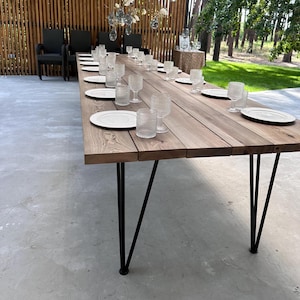 Könnte beinhalten: Ein langer rechteckiger Esstisch mit einer Oberfläche aus natürlichen Holzplanken und schwarzen Metall-Haarnadelbeinen. Der Tisch ist mit weißen Keramiktellern und klaren, strukturierten Glasbechern gedeckt, die in Reihen angeordnet sind.