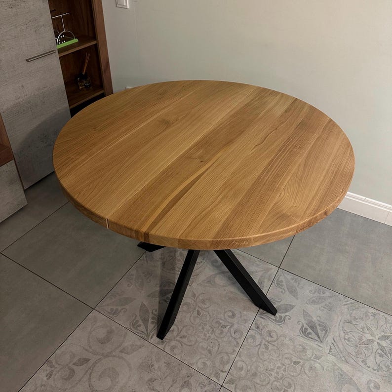 K&ouml;nnte beinhalten: Ein runder Esstisch mit einer Tischplatte aus hellbraunem Holz mit sichtbarer Maserung und glatter Oberfl&auml;che. Der Tisch steht auf einem schwarzen Metallgestell, das aus vier schr&auml;g verlaufenden Beinen besteht, die in der Mitte zusammenlaufen.