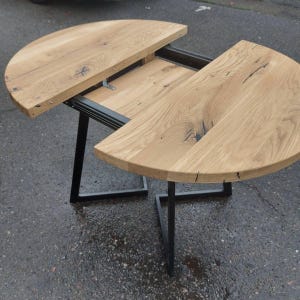 May include: An extendable table featuring two semi-circular light brown wood grain surfaces with visible knots and dark fissures. The table is supported by a black metal frame with angular legs and a sliding mechanism that separates the two halves.