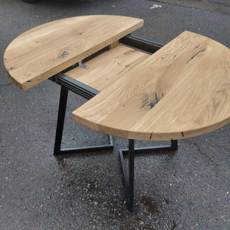 May include: An extendable table featuring two semi-circular light brown wood grain surfaces with visible knots and dark fissures. The table is supported by a black metal frame with angular legs and a sliding mechanism that separates the two halves.