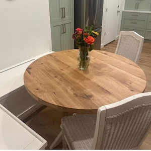 May include: A round light brown wooden dining table with a visible natural grain pattern. A clear glass vase sits in the center of the table, containing a bouquet of orange, red, and yellow flowers with green stems and leaves.
