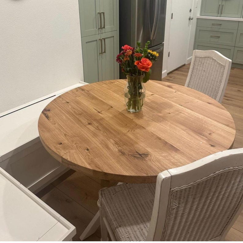 May include: A round light brown wooden dining table with a visible natural grain pattern. A clear glass vase sits in the center of the table, containing a bouquet of orange, red, and yellow flowers with green stems and leaves.