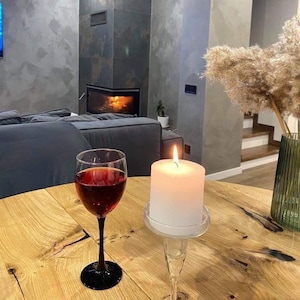 May include: A clear glass wine goblet filled with deep red wine and a white pillar candle in a clear glass holder sit on a light-colored wooden table. The wine glass has a black stem and base, while the candle emits a warm, glowing flame.