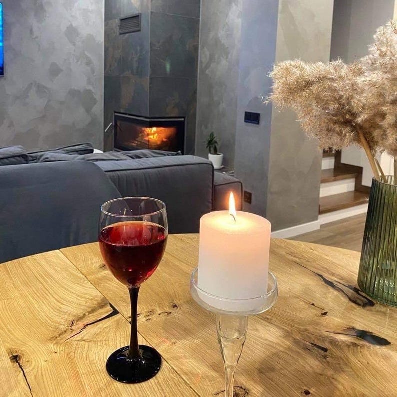 May include: A clear glass wine goblet filled with deep red wine and a white pillar candle in a clear glass holder sit on a light-colored wooden table. The wine glass has a black stem and base, while the candle emits a warm, glowing flame.