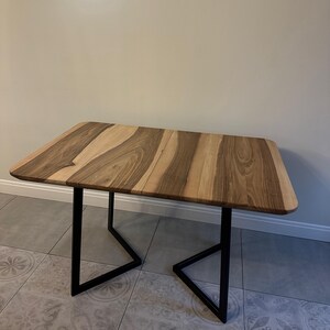 K&ouml;nnte beinhalten: Ein rechteckiger Holztisch mit einem nat&uuml;rlichen Holzmaserungsmuster in Braun- und Beiget&ouml;nen. Der Tisch hat schwarze Metallbeine. Der Tisch ist etwa 100 cm lang und 60 cm breit.