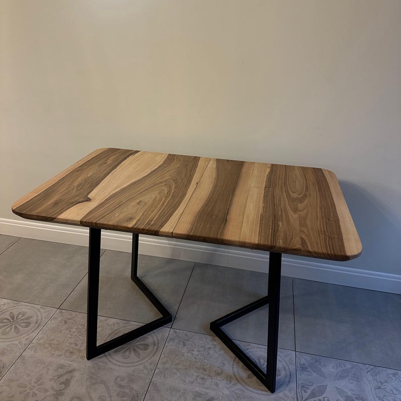 K&ouml;nnte beinhalten: Ein rechteckiger Holztisch mit einem nat&uuml;rlichen Holzmaserungsmuster in Braun- und Beiget&ouml;nen. Der Tisch hat schwarze Metallbeine. Der Tisch ist etwa 100 cm lang und 60 cm breit.