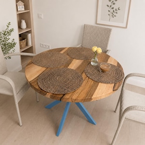 May include: A round wooden dining table with a natural grain finish and a bright blue metal base. The table is set with three circular woven brown placemats, a small clear glass vase containing two yellow flowers, and a clear glass bowl with liquid.