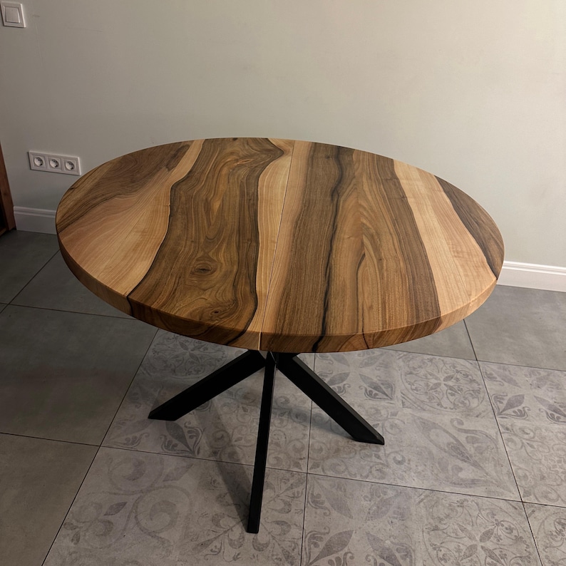 May include: A round dining table featuring a natural wood grain tabletop with light and dark brown tones. The table is supported by a black metal base consisting of four angled legs that meet in the center, providing a stable foundation for the top.