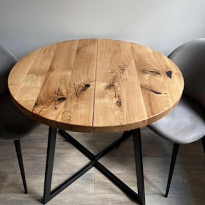 May include: A round wooden dining table with a natural light brown grain, visible knots, and a central seam. The tabletop is supported by a black metal base featuring an X-shaped frame design. Two grey upholstered chairs are positioned on each side.