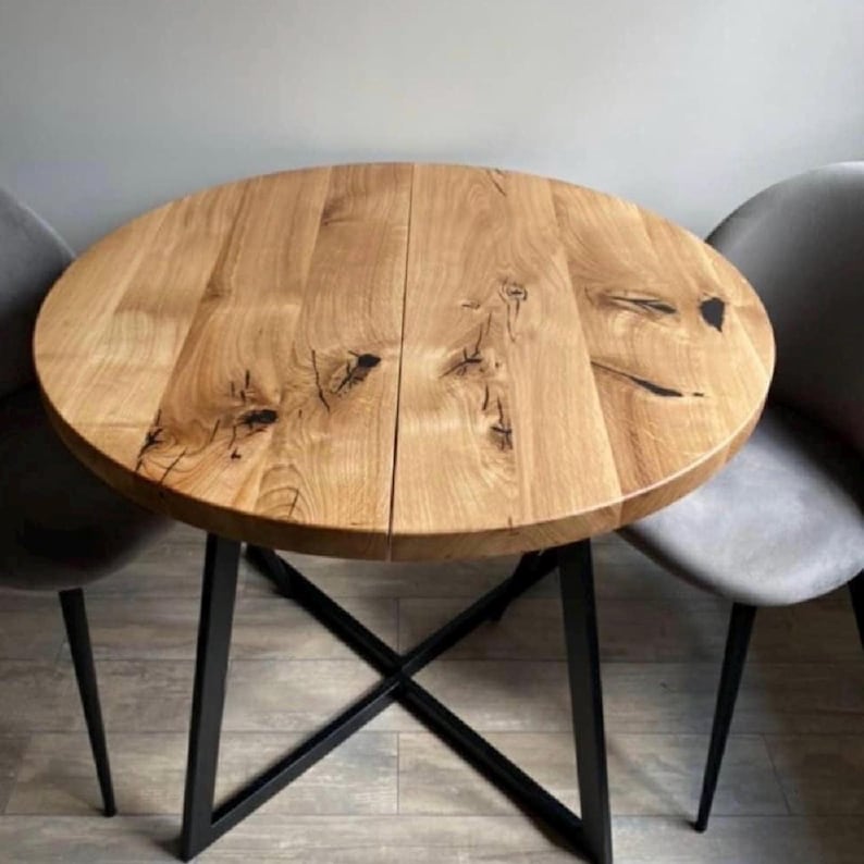May include: A round wooden dining table with a natural light brown grain, visible knots, and a central seam. The tabletop is supported by a black metal base featuring an X-shaped frame design. Two grey upholstered chairs are positioned on each side.