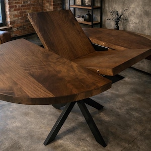 Può includere: Tavolo da pranzo allungabile marrone scuro, con piano rotondo e base nera a forma di stella. Il tavolo è mostrato parzialmente esteso, rivelando il meccanismo interno. La venatura del legno è visibile.
