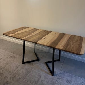 K&ouml;nnte beinhalten: Ein rechteckiger Holztisch mit nat&uuml;rlicher Holzmaserung. Die Tischplatte wird von schwarzen Metallbeinen getragen. Der Tisch ist ca. 183 cm lang und 91 cm breit und eignet sich zum Essen oder Arbeiten.