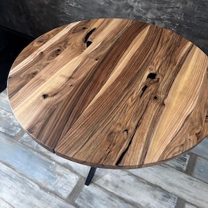May include: A round table featuring a natural wood grain surface with visible knots and dark brown streaks. The tabletop is constructed from joined wood planks, showcasing a varied light and dark brown color palette with a smooth finish.