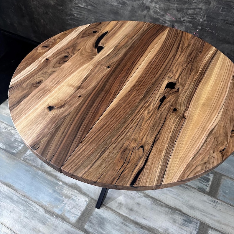 May include: A round table featuring a natural wood grain surface with visible knots and dark brown streaks. The tabletop is constructed from joined wood planks, showcasing a varied light and dark brown color palette with a smooth finish.