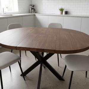 Puede incluir: Una mesa de comedor redonda de madera con veta visible y una hoja central extensible, apoyada sobre una base de metal negro en forma de X. Cuatro sillas con asientos tapizados en gris claro y patas de metal negro rodean la mesa.