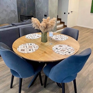 May include: A round light wood dining table with four blue velvet chairs. The table features four circular placemats with a geometric pattern in black, white, and tan. A green glass vase filled with beige pampas grass sits in the center.