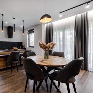 May include: A round wooden dining table with a natural grain finish and a black metal base, surrounded by four dark gray upholstered chairs. A glass vase filled with dried pampas grass sits in the center of the table in a dining room.