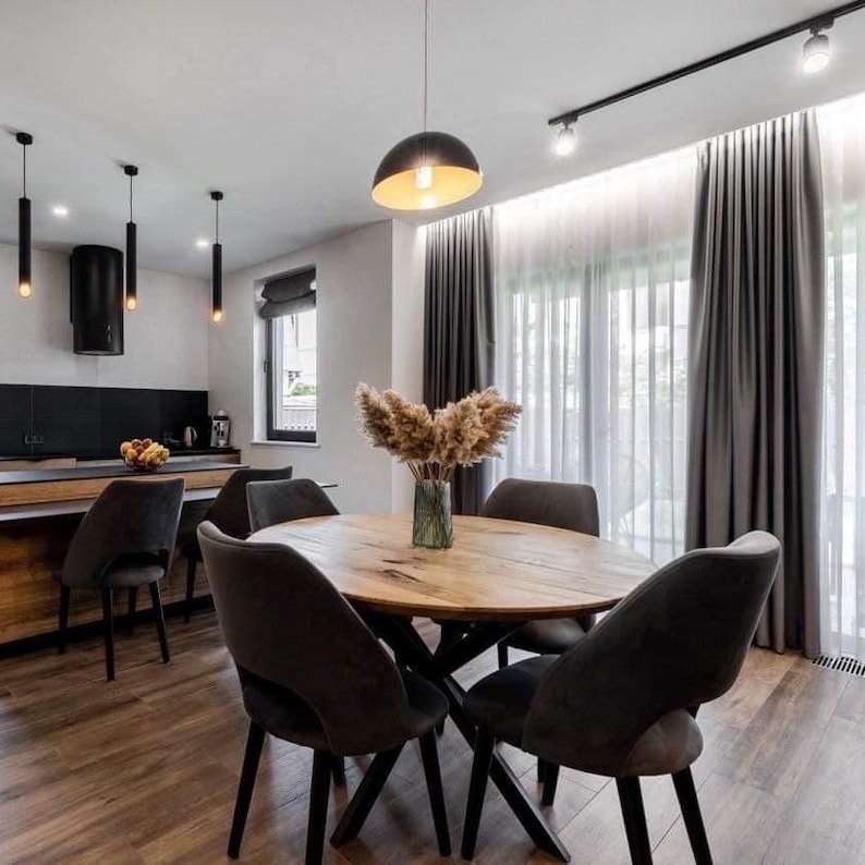 May include: A round wooden dining table with a natural grain finish and a black metal base, surrounded by four dark gray upholstered chairs. A glass vase filled with dried pampas grass sits in the center of the table in a dining room.