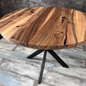 May include: A round dining table featuring a natural wood grain top with visible knots and dark brown streaks. The tabletop is constructed from multiple joined planks and is supported by a black metal base with four angled legs meeting at a center point.