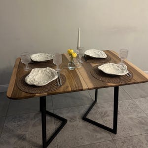 K&ouml;nnte beinhalten: Ein Holztisch f&uuml;r vier Personen. Der Tisch hat eine nat&uuml;rliche Holzmaserung und schwarze Metallbeine. Jedes Gedeck umfasst einen strukturierten wei&szlig;en Teller, einen gewebten Platzdeckchen, ein Wasserglas und Besteck. Eine kleine Vase mit gelben Blumen und eine Kerze vervollst&auml;ndigen die Dekoration.