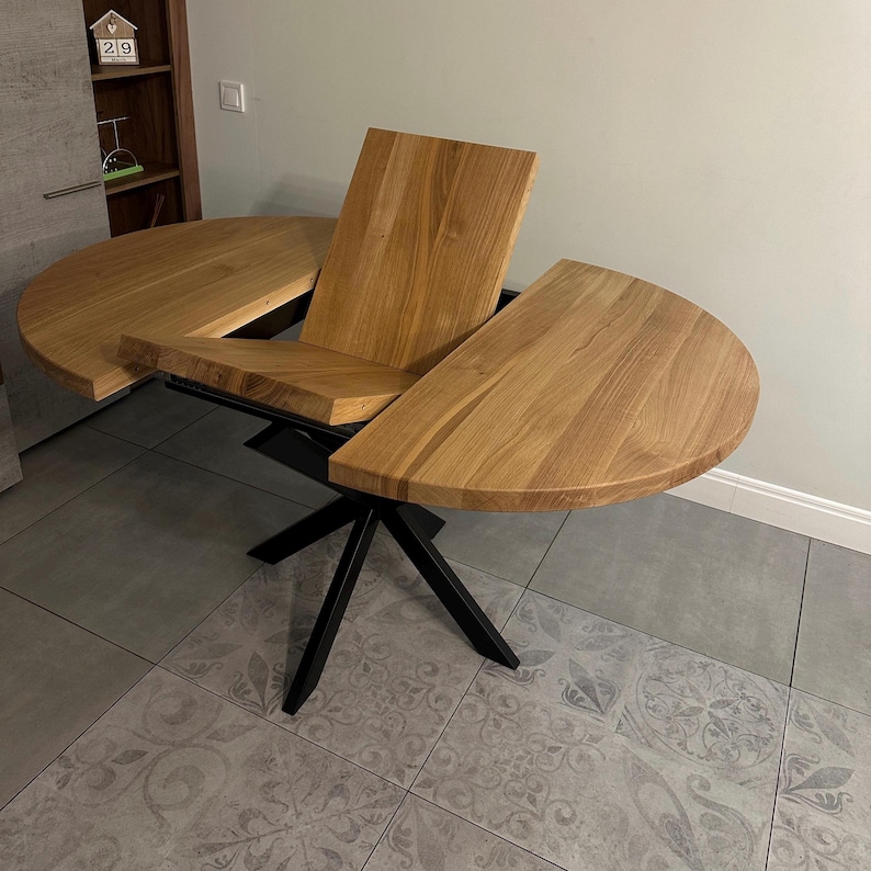 K&ouml;nnte beinhalten: Ein runder Esstisch aus Holz mit einer nat&uuml;rlichen hellbraunen Holzmaserung. Der Tisch verf&uuml;gt &uuml;ber einen ausziehbaren Mittelteil und wird von einem schwarzen Metallgestell mit vier schr&auml;gen Beinen getragen.
