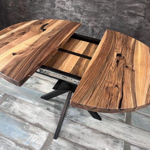 May include: A round wooden dining table with a natural wood grain finish. The table has a split top that can be extended. The base is made of black metal. The table is approximately 60 inches in diameter when closed.