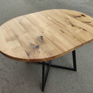 May include: An oval dining table featuring a light brown natural wood grain top with visible dark knots and cracks. The tabletop is supported by a black metal base constructed with intersecting legs that form an X-shaped support structure.