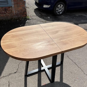 Pode incluir: Uma mesa de jantar oval com um tampo de madeira castanho-claro com veios visíveis e uma costura central, apoiada por uma base de metal preto com um design de estrutura em forma de X. A superfície da mesa apresenta uma textura de madeira natural.