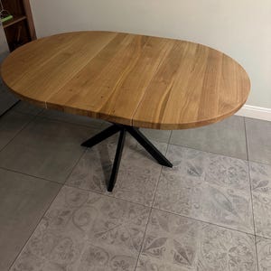 K&ouml;nnte beinhalten: Ein ovaler Esstisch mit einer Tischplatte aus hellbraunem Holz mit sichtbarer Maserung und Fugen, die auf ein ausziehbares Design hinweisen. Der Tisch steht auf einem zentralen schwarzen Metallgestell mit vier schr&auml;g zulaufenden Beinen.
