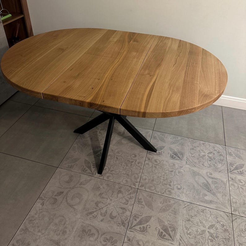 K&ouml;nnte beinhalten: Ein ovaler Esstisch mit einer Tischplatte aus hellbraunem Holz mit sichtbarer Maserung und Fugen, die auf ein ausziehbares Design hinweisen. Der Tisch steht auf einem zentralen schwarzen Metallgestell mit vier schr&auml;g zulaufenden Beinen.