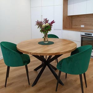 May include: A round wooden dining table with a dark metal base, surrounded by three green velvet chairs. A vase of flowers sits on the table's center, atop a green placemat. The table is in a kitchen setting with white cabinets.