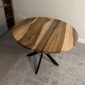 May include: A round dining table featuring a natural wood grain tabletop with alternating light and dark brown tones. The table is supported by a black metal base consisting of four angled legs that meet in the center beneath the circular top.