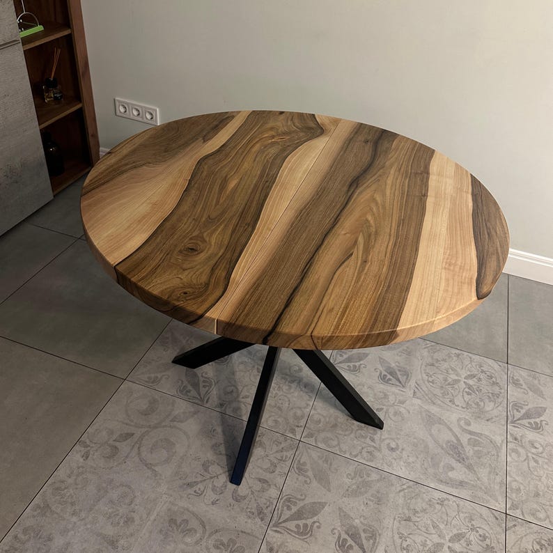May include: A round dining table featuring a natural wood grain tabletop with alternating light and dark brown tones. The table is supported by a black metal base consisting of four angled legs that meet in the center beneath the circular top.