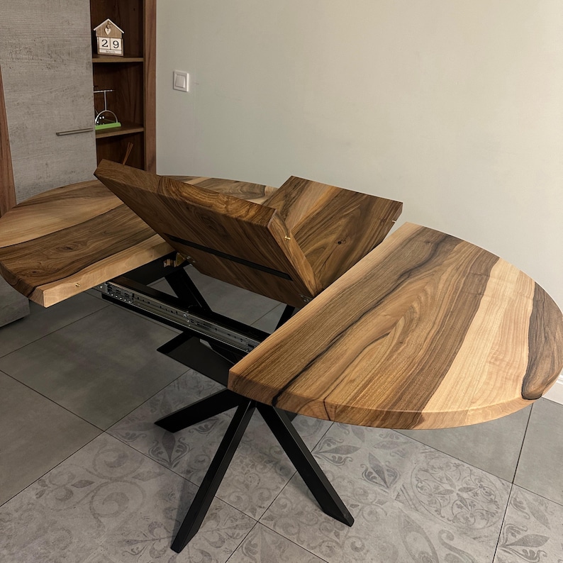May include: An extendable dining table featuring a natural wood grain surface with dark and light brown tones. The table has a central folding leaf mechanism and is supported by a black metal base with angled legs and visible sliding tracks.