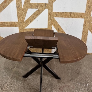 Pode incluir: Uma mesa de jantar redonda de madeira com acabamento castanho escuro e veio de madeira visível. A mesa possui um mecanismo extensível com uma secção central e uma base de metal preto com quatro pernas cruzadas em forma de X.