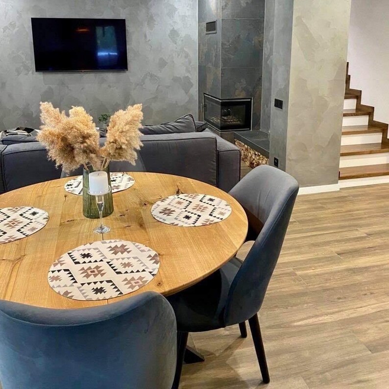 May include: A round light wood dining table with four blue upholstered chairs. The table features four circular placemats with a geometric pattern in brown, black, and white. A glass vase filled with dried pampas grass sits in the center.