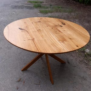May include: A round wooden table with a natural finish. The tabletop is made of light brown wood planks and is supported by a three-legged base. The table is approximately 122 cm in diameter.
