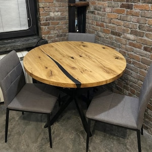 May include: A round wooden dining table with a black resin inlay, supported by a black metal base. Two gray upholstered chairs are positioned around the table. The table is set against a brick wall.