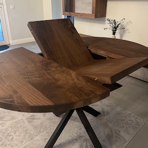 Peut inclure: Une table de salle &agrave; manger extensible brun fonc&eacute; au design unique. Le plateau est en bois et pr&eacute;sente un motif de grain visible. La table a un pi&egrave;tement en m&eacute;tal noir angulaire. La table est partiellement d&eacute;ploy&eacute;e, montrant le m&eacute;canisme de pliage.