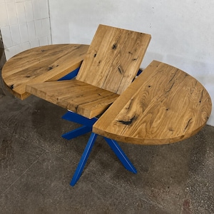 May include: A wooden dining table with a unique design. The tabletop is made of light brown wood with visible grain and a semi-circular shape. The base is a vibrant blue, with angled legs. The table is designed to expand.