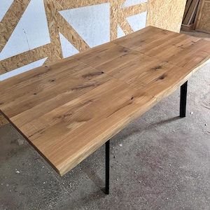 May include: A rectangular dining table featuring a natural light brown wood grain tabletop with visible knots and a live edge. The table is supported by four thin, vertical black metal legs positioned at the corners of the wooden surface.